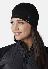 Gorro negro con costuras planas y un pequeño logotipo de personaje en el lateral, usado con una chaqueta negra con cremallera que presenta secciones acolchadas.