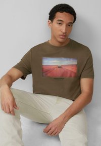 Olijfgroene t-shirt met een vooropdruk van een auto op een weg, met daaronder de tekst "MOVING STORIES". Witte, gestructureerde stoffen broek.