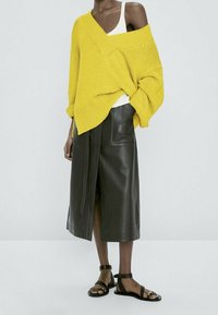 Pull en maille jaune oversize à col bateau, associé à une jupe midi en cuir foncé avec une fente frontale, et des sandales noires.