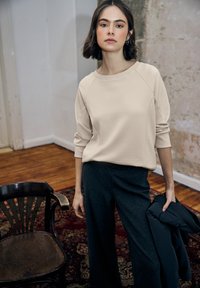 Sudadera beige con mangas raglán, combinada con pantalones oscuros de rayas finas. Modelo sosteniendo una chaqueta negra, de pie sobre un suelo de madera.