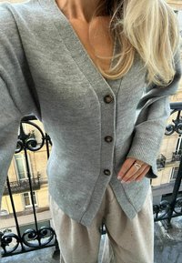 Světle šedý pletený kardigan s V-výstřihem, pěti tmavými knoflíky, žebrovanými manžetami a slim fit střihem. V kombinaci se světlými texturovanými kalhotami.