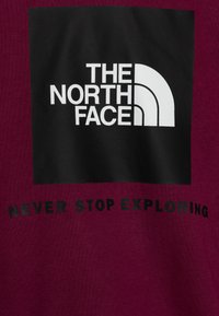 Bordowa bluza z czarnym prostokątnym logo z napisem "THE NORTH FACE" oraz białym półokręgiem. Poniżej napis "NEVER STOP EXPLORING."