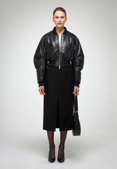 Giacca bomber nera in pelle con maniche voluminose sopra una gonna nera al ginocchio. L'outfit è abbinato a scarpe con tacco alto nere e a una borsa.