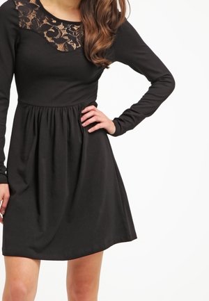 Femme portant une robe noire à manches longues avec un empiècement en dentelle, main sur la hanche, sur fond blanc.