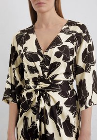 Floral dress in cream with black flower print, featuring a deep V-neck and knotted waist. Made of smooth fabric with wide sleeves.