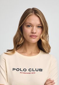 Kurzärmliges, off-white T-Shirt mit dem gestickten Schriftzug "POLO CLUB ESTABLISHED 2012" in marineblauen und roten Buchstaben über der Brust. Weicher Stoff.