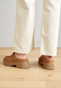 Zoccoli in suede marrone con una spessa suola piatta di un marrone chiaro. Le scarpe hanno un design slip-on e una texture liscia, con dettagli di cucitura sottili.
