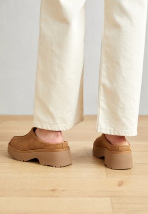 Zuecos de ante marrón con una gruesa suela de plataforma de color marrón claro. Los zapatos tienen un diseño sin cordones y una textura suave, con detalles de costuras sutiles.