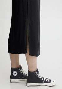 Čierna, zvrásnená midi sukňa s bočným rozparkom, v kombinácii s čiernymi vysokými teniskami Converse s bielymi akcentmi.