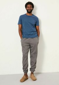 Blaues Baumwoll-T-Shirt mit Fronttasche, kombiniert mit grauen Sweatpants. Das Modell trägt braune Slipper mit einer abgerundeten Spitze und seitlichem Schnallenverschluss.