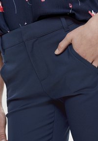 Marinblå skräddarsydda byxor i slät tyg, med en diskret sidoficka och en slim fit-design. Händerna placerade i framfickan.