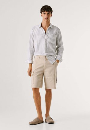 Noor mees, kes kannab valget triibulist nööbitavat särki, beeže vööpükse ja sobivaid beeže libisevaid jalatseid, seisab ühe käega taskus.