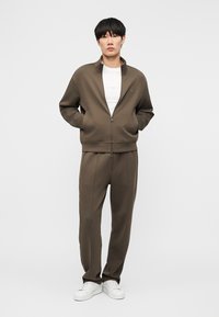 Νεαρός άνδρας στέκεται με τα χέρια στις τσέπες, φορώντας χακί καφέ μπουφάν με φερμουάρ και ταιριαστό παντελόνι, λευκά αθλητικά παπούτσια και λευκό μπλουζάκι.