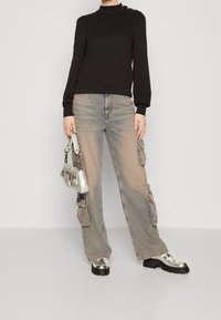 Schwarzes Oberteil mit langen Ärmeln und Schulterknöpfen, kombiniert mit ausgewaschenen, im Cargo-Stil gehaltenen Jeans mit Seitentaschen. Silberschultertasche und glänzende Schuhe.