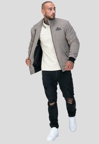 Veste bomber à motif pied-de-poule avec col côtelé, bord noir et doublure intérieure beige. Portée avec un jean noir déchiré et des sneakers blanches.