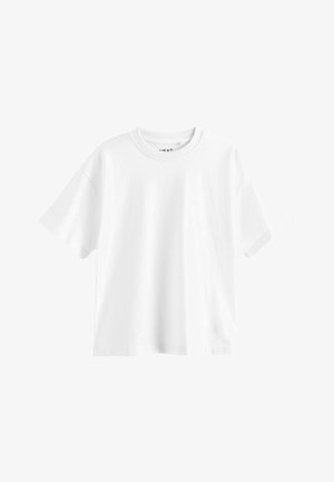Ung dreng går iført en løs hvid t-shirt, lyseblå cargobukser og grå sneakers mod en ensfarvet lys baggrund.