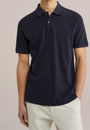 Mężczyzna w granatowej koszulce polo z krótkim rękawem, noszący luźno włożone beżowe spodnie, stojący na jednolitym beżowym tle.