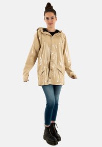 Impermeable beige y brillante con capucha, botones de presión y bolsillos laterales. Se lleva sobre una camiseta oscura, combinado con jeans azules y botas negras con cordones.