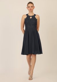 Donker marineblauwe cocktailjurk met een nauwsluitend kantengedeelte, halterhalslijn en geplooide chiffon rok. Bevat een sleuf met sleutelgat en glanzende sandalen.