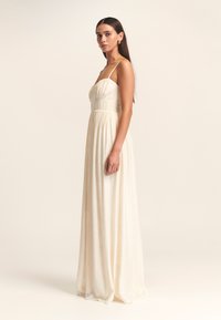 Lang, vloeiend jurkje van off-white stof; heeft dunne spaghettibandjes, een aansluitend lijfje en een geraffineerde taille die leidt naar een volle rok.