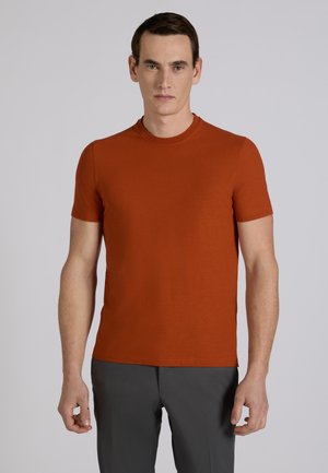 Mann trägt ein rostfarbenes T-Shirt mit kurzen Ärmeln und Rundhalsausschnitt sowie dunkelgraue Hose, steht vor einem einfarbigen hellgrauen Hintergrund.