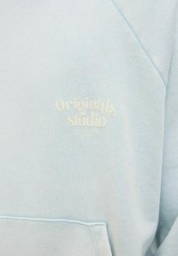 Light blue sweatshirt made of textured fabric, featuring a kangaroo pocket and the words "Originals Studio" in subtle white lettering.