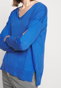 Pull en laine bleu avec un col en V, des manches longues et une coupe ample. Le tissu texturé présente des fentes latérales à l'ourlet pour un détail supplémentaire.