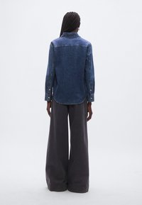 Farkkupaita, jossa on suora malli, pitkät hihat ja terävä kaulus. Tummansininen väri, vaaleat saumat ja kaartuva helma. Housut ovat leveälahkeiset ja harmaat.