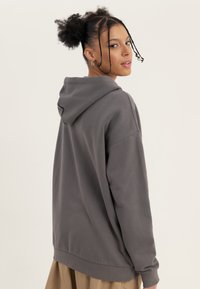 Mladá žena s dvěma afro copánky, nosící kruhové náušnice a šedou mikinu s kapucí, otočená mírně dozadu a dívající se přes rameno.