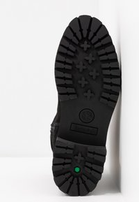 Semelle d'une botte Timberland noire avec une semelle profonde et un détail hexagonal vert près du talon, sur fond blanc.