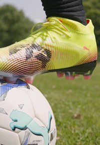 Lys gul fotballsko med teksturert overflate og svarte striper, plassert på en hvit og blå mønstrete fotball på gress.