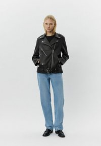Chaqueta de motorista de piel negra con herrajes plateados, cremallera asimétrica, bolsillos laterales y cintura con cinturón; combinada con jeans azul claros y zapatos negros.