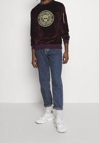 Sweatshirt in velluto borgogna con grafica di un leone, abbinato a jeans blu a righe. Sneakers bianche completano l'outfit.