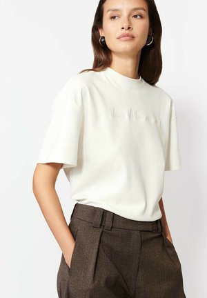 Biała krótka koszulka z krótkim rękawem, o wysokim dekolcie i haftowanym napisem. Noszona z brązowymi, fakturowanymi spodniami o wysokim stanie.