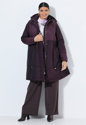 Femme souriante portant un manteau matelassé violet foncé avec capuche, un pantalon large et des bottes à talons, debout devant un fond blanc uni.