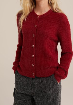 Kvinde iført en blød, rød cardigan med knapper kombineret med mørkegrå teksturerede bukser, hænderne i lommerne, foran en neutral baggrund.