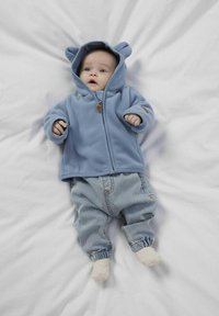 Felpa in pile azzurro chiaro con orecchie da orso, zip frontale e patch sul petto, abbinata a pantaloni di jeans azzurro chiaro, su sfondo bianco.