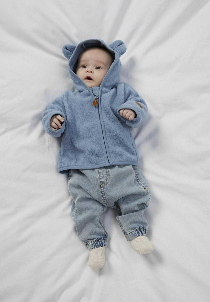 Felpa in pile azzurro chiaro con orecchie da orso, zip frontale e patch sul petto, abbinata a pantaloni di jeans azzurro chiaro, su sfondo bianco.