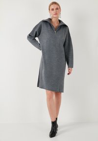 Graues Strickkleid mit Reißverschlusskragen, lockerer Passform, langen Ärmeln, seitlichen Schlitzen und glatter Textur, kombiniert mit schwarzen Knöchelstiefeln.