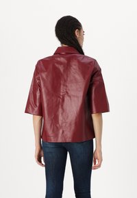 Burgunderfarget faux skinnjakke med krage, korte ermer og glatt tekstur, kombinert med tettsittende blå jeans.