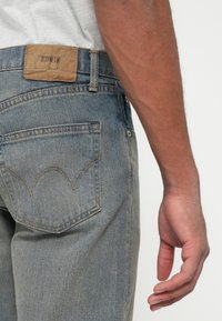 Jeans din denim albastru estompat, cu un label din piele EDWIN pe talie la spate și detalii vizibile de cusătură pe buzunarele din spate.