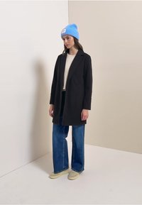 Zwarte op maat gemaakte jas met een notched revers, gecombineerd met een beige top, brede blauwe jeans en een lichtblauwe gebreide muts. Casual schoenen zichtbaar.