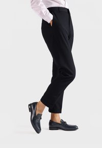 Pantaloni neri su misura con tasche, indossati con mocassini in pelle blu scuro con dettagli metallici, presentati su uno sfondo chiaro.