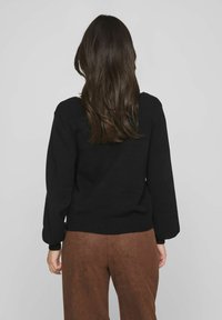 Suéter de punto negro con una textura suave, que presenta un amplio escote redondo, mangas largas y un dobladillo acanalado, combinado con pantalones de ante marrón.