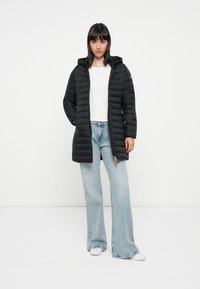 Zwarte gewatteerde jas met een capuchon, rits aan de voorkant en ribgebreide manchetten; gecombineerd met lichtblauwe flared jeans en witte sneakers. Opvallende oranje ritssluiting.