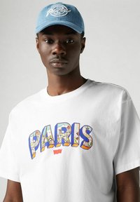 Camiseta blanca de algodón con un gráfico colorido de "PARÍS" que presenta edificios; combinada con una gorra de denim azul claro bordada con "París."