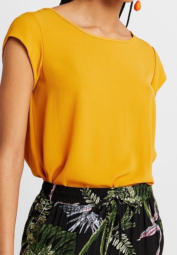 Donna che indossa una maglietta corta gialla senape infilata in pantaloni neri a vita alta con stampa di foglie tropicali verdi e rosa.