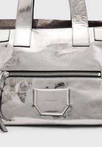 Sølvmetallic tote bag med tekstureret læder, dobbelte håndtag, en lynlåslomme og et logo patch. Har et minimalistisk design.