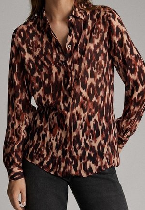 Woman wearing a long-sleeve, button-up blouse with brown, black, and beige abstract animal print tucked into black pants.