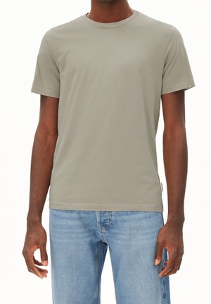 Dunkelhäutige Person trägt ein schlichtes hellgraues kurzärmeliges T-Shirt und hellblaue Jeans, dargestellt vom Hals bis zu den Hüften.